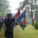 Grupa mundurowych w deszczu salutuje przy flagach. Tło z zielonymi drzewami. Centralnie unoszona biało-czerwona flaga.