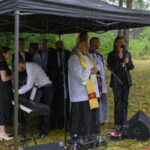 Grupa ludzi pod czarnym namiotem, podczas deszczu. Na pierwszym planie duchowny w stroju liturgicznym. Tło stanowi zielona roślinność.