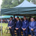 Grupa osób w garniturach stoi pod zielonym namiotem. Pada deszcz, a w tle widać zielone drzewa i kolejny namiot z ludźmi. Po lewej stoi kilka pustych krzeseł na trawie.