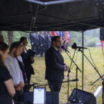 Pod namiotem grupa ludzi uczestniczy w ceremonii przy mikrofonie. W tle widoczne są drzewa i flagi Polski. Pada deszcz.