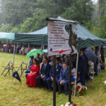 Obraz przedstawia uroczystość plenerową pod zielonymi namiotami w deszczu. Ludzie siedzą na krzesłach na trawie, z kwiatami wokół. W centrum widnieje tablica upamiętniająca historyczne wydarzenie.