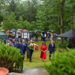 Grupa ludzi z kwiatami na pewno w uroczystości plenerowej. Zielona trawa, drzewa i namioty w tle. Parasole w różnych kolorach chronią przed deszczem.