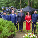 Grupa ludzi stoi na trawniku podczas uroczystości w parku. Mają na sobie eleganckie ubrania; jest deszczowo, widoczne parasole. W tle gęsta zieleń i namiot.