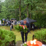 Mężczyzna w czarnym garniturze składa wieniec na tle zielonego lasu. Grupa osób z parasolami w tle. Czerwone kwiaty z przodu.