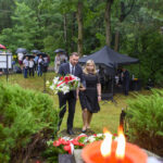 Dwójka ludzi składa wiązankę kwiatów przed pomnikiem. W tle zielony las, uczestnicy z parasolkami. Pierwszy plan to płomień świecy.