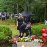 Mężczyzna i kobieta składają kwiaty na grobie, w tle zielone drzewa. Widać czarne parasole i czerwone kwiaty. Tło stanowi las.