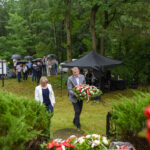 Na zdjęciu widoczni są ludzie pod parasolami w lesie podczas ceremonii. Mężczyzna niesie wieniec z biało-czerwonymi kwiatami. W tle namiot i drzewa.