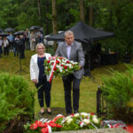 Para składa wieniec na tle zieleni. Ludzie z parasolami stoją w oddali. Czarny namiot widoczny po prawej stronie.