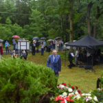 Grupa ludzi z parasolkami stoi na trawie wśród drzew. Na pierwszym planie kwiaty, z tyłu zielony i czarny namiot. Tło to bujna zieleń i deszczowa atmosfera.