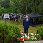 Mężczyzna w garniturze stoi przed czerwono-białymi kwiatami. W tle widoczne są drzewa, ludzie z parasolami i czarne namioty. Dominuje zielona sceneria.