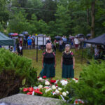 Dwie starsze kobiety w ludowych strojach stoją przed wieńcami. W tle zebrani ludzie z parasolami. Scena na tle zieleni.