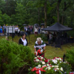 Ludzie składają wieńce w zielonym parku. W tle namioty i tłum pod parasolami. Atmosfera deszczowa, dominują zielenie i czerwienie kwiatów.