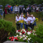 Grupa osób w białych koszulkach z kwiatami stoi przed wieńcami. Tło zielone, ludzi osłaniają kolorowe parasole. Atmosfera poważna.