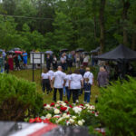 Grupa ludzi pod parasolami na tle zielonego lasu. Namioty w ciemnych kolorach. Kwiaty w jasnych barwach na pierwszym planie.