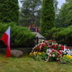 Nagrobek z krzyżem otoczony zielenią, kwiaty biało-czerwone. Polska flaga z lewej, tło to drzewa i trawa.
