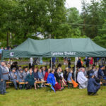 Na zdjęciu jest zielony namiot z napisem Gmina Dobre. Pod namiotem siedzą ludzie. W tle widać drzewa i rowery.