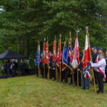 Grupa osób w paradnych strojach trzyma kolorowe sztandary na tle zielonego lasu. Obok czarny namiot i mężczyźni w mundurach. Trawa jest wilgotna, a niebo pochmurne.