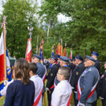Grupa osób w mundurach trzyma sztandary w parku. Ubrani w ciemne garnitury, otoczeni zielenią drzew. Czerwono-białe flagi dominują.
