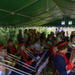 Orkiestra dęta w granatowych mundurach z czerwonymi akcentami gra pod zielonym namiotem. Publiczność siedzi w tle. Na zewnątrz zielone krzewy.