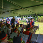 Orkiestra w kolorowych mundurach gra pod zielonym namiotem. Dyrygent w czerwonym płaszczu. Na tle zieleń i publiczność z parasolami.