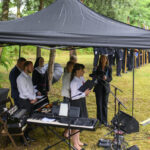 Grupa ludzi pod czarnym namiotem z mikrofonem i instrumentami, w tle zielone drzewa i stojąca straż honorowa.