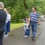 Na zdjęciu widzimy ludzi stojących w kolejce na mokrej drodze. Mężczyzna z dziewczynką trzymającą pluszaka idą obok. W tle zielone drzewa i zaparkowane samochody.