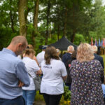 Grupa ludzi w parku uczestniczy w ceremonii. W tle flagi i namiot, otoczenie jest zielone. Dorośli i dzieci w odświętnych strojach.