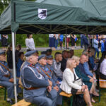 Grupa ludzi pod zielonym namiotem, część w mundurach, siedzi na ławkach. W tle widać widzów na trawie. Pogoda pochmurna.