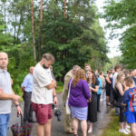 Grupa ludzi stoi na leśnej ścieżce, otoczona zielenią drzew. Ubrani są w różnorodne stroje od casualowych po eleganckie. Tło stanowią bujne drzewa i roślinność.