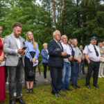 Grupa osób stoi na trawie w parkowym otoczeniu. Ubrani są elegancko, a w tle widać drzewa. Atmosfera jest formalna i spokojna.
