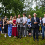 Grupa ludzi stoi na trawie, w eleganckich i casualowych strojach, na tle zieleni drzew. Część osób trzyma dokumenty. Pogoda jest pochmurna.