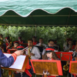 Orkiestra w czerwonych mundurach gra pod zielonym namiotem. Dyrygent kieruje zespołem. W tle zielone krzewy.