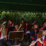 Orkiestra w czerwono-niebieskich mundurach gra pod zielonym namiotem. Tło to bujna zieleń. Instrumenty to głównie dęte i saksofony.