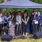 Grupa ludzi pod czarnym namiotem na tle zieleni. Osoby są elegancko ubrane, część trzyma mikrofony i dokumenty. Tło to drzewa i samochód.