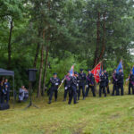 Grupa strażaków maszeruje z flagami po zielonym trawniku, otoczonym drzewami. Na lewo stoi namiot z widownią. Na trawie leżą kwiaty.