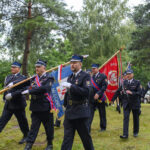 Strażacy w mundurach niosą flagi na zielonym tle drzew. Dominujące kolory to niebieski, czerwony i zielony. Ceremonialny marsz.
