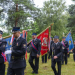 Na zdjęciu widać grupę strażaków w paradnych mundurach niosących sztandary. Tło stanowią zielone drzewa i trawa. Kolory dominujące to czerń mundurów oraz czerwony i niebieski sztandarów.