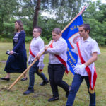 Na zdjęciu grupa młodych osób w białych koszulach niesie niebieską flagę. Tło stanowi zielona trawa i drzewa. Atmosfera jest uroczysta, a kolory żywe.