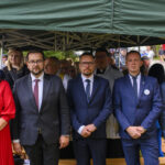 Pod zielonym namiotem stoi grupa elegancko ubranych osób. Mężczyźni w garniturach, kobieta w czerwonej sukience. W tle widać uczestników wydarzenia.