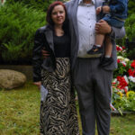 Grupa osób pozuje na trawie, w tle zieleń i kwiaty. Mężczyzna trzyma dziecko. Kobieta w czarno-białej spódnicy.