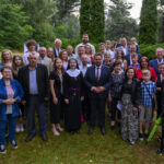 Grupa ludzi pozuje do zdjęcia na tle zielonego lasu. Mają różnorodne stroje, w tym eleganckie garnitury i sukienki. W oddali widać niebieski namiot oraz flagi.