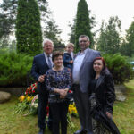 Grupa pięciu osób stoi na tle zielonych drzew i krzewów. Mężczyźni mają na sobie garnitury, a kobiety eleganckie ubrania. Widać kwiaty i znicze na trawie.