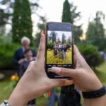 Telefon w rękach fotografuje grupę osób na tle zieleni. W tle widać drzewa i flagi. Kolory są jasne, dominują zieleń i biel.