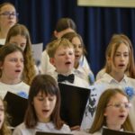 Grupa dzieci ubranych na biało śpiewa. Mają czarne teczki z nutami i niebieskie kwiaty. Tło stanowi ciemna zasłona.