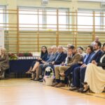 Grupa ludzi siedzi w sali gimnastycznej na drewnianej podłodze. Z lewej strony widnieje baner z napisem Mazowsze. W tle drabinki gimnastyczne i okna.