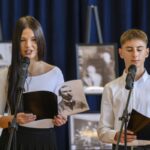 Na tle granatowej zasłony dwoje młodych ludzi w białych koszulach recytuje poezję z mikrofonami. Trzymają czarno-białe zdjęcia znanych postaci. W tle widoczne są rozmyte obrazy na sztalugach.