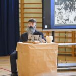 Mężczyzna w przebraniu siedzi przy stole pokrytym papierem. Na ekranie za nim widać czarno-białą fotografię grupową. Tło jest granatowe z drewnianymi elementami.