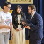 Trzy osoby stoją w jasnym pokoju; kobieta w białej koszulce, mężczyzna w garniturze wręcza jej dokument. Tło ciemnoniebieskie z kwiatami.