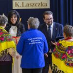 Na zdjęciu dwie kobiety w żółtych chustach i kobieta oraz mężczyzna w eleganckich strojach. Tło jest ciemnoniebieskie z napisem. Widać czerwone kwiaty w wazonach.