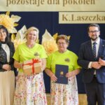 Na zdjęciu cztery osoby na tle dekoracji z żółtych i białych kwiatów. Dwie kobiety w zielonych koszulkach trzymają prezenty. Mężczyzna i kobieta są elegancko ubrani.